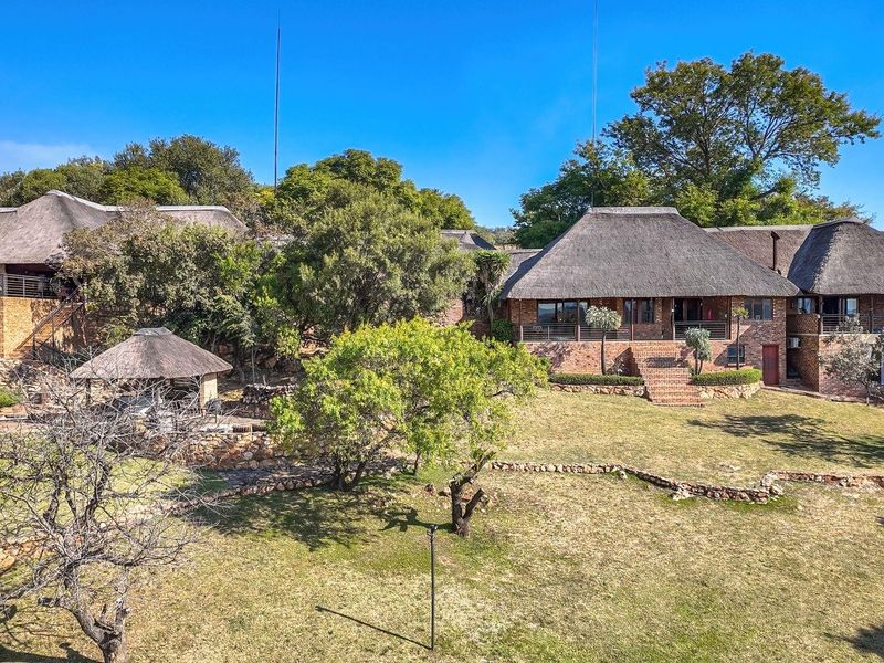 An elevated view of a spacious farm property featuring multiple thatch-roofed buildings, face brick construction, stone retaining walls, and a landscaped garden with a pool.