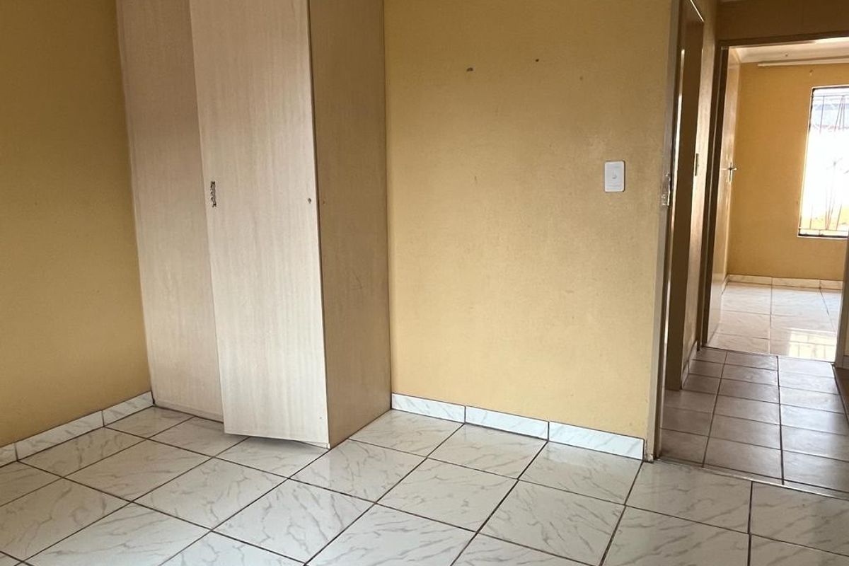 An empty bedroom featuring light-colored ceramic floor tiles and a built-in wardrobe, with a view into the adjacent hallway.