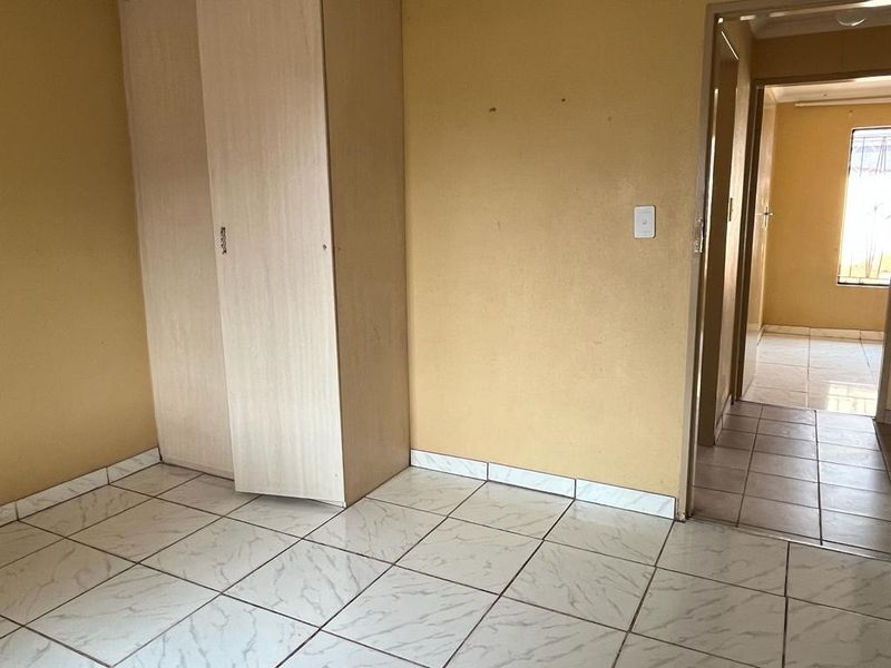 An empty bedroom featuring light-colored ceramic floor tiles and a built-in wardrobe, with a view into the adjacent hallway.