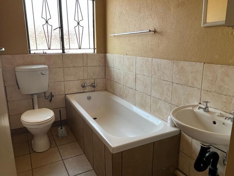 A functional bathroom featuring a bathtub, pedestal basin, and toilet, with tiled splashbacks and security bars on the window.