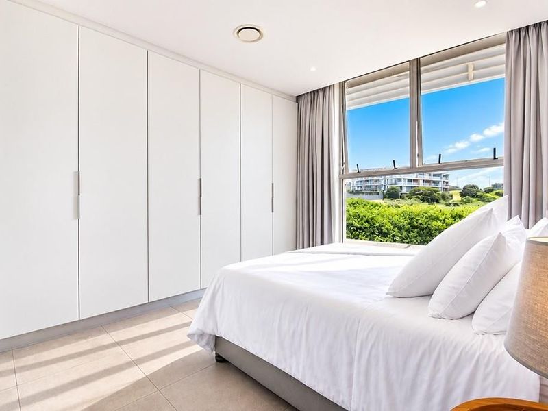 A bright, modern bedroom featuring extensive built-in floor-to-ceiling wardrobes, tiled flooring, and large windows offering pleasant exterior views.
