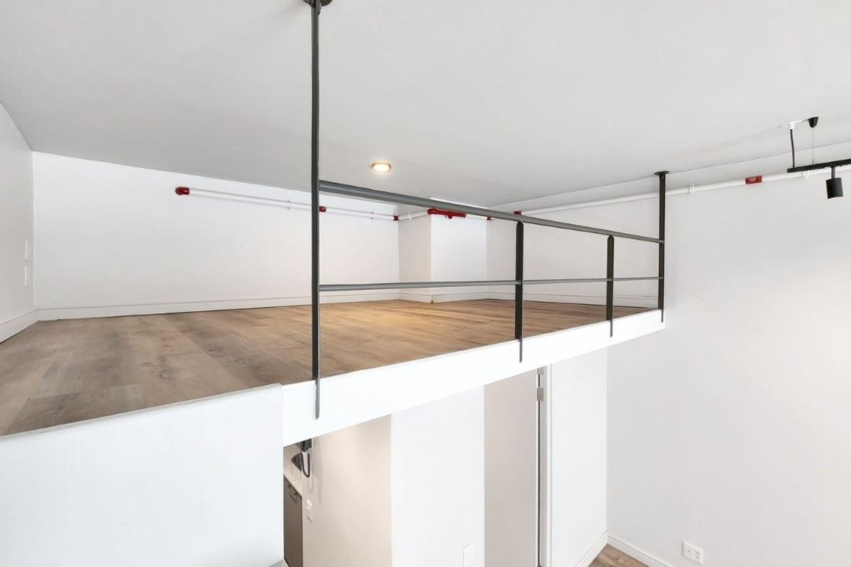 A bright and modern mezzanine bedroom area featuring wood-look laminate flooring, a sleek metal safety railing, and exposed industrial-style fire sprinkler piping.