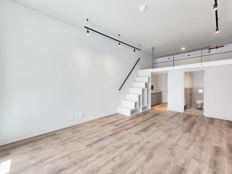 Modern open-plan apartment featuring light laminate flooring, a mezzanine level with metal railing, built-in staircase storage, and track lighting.