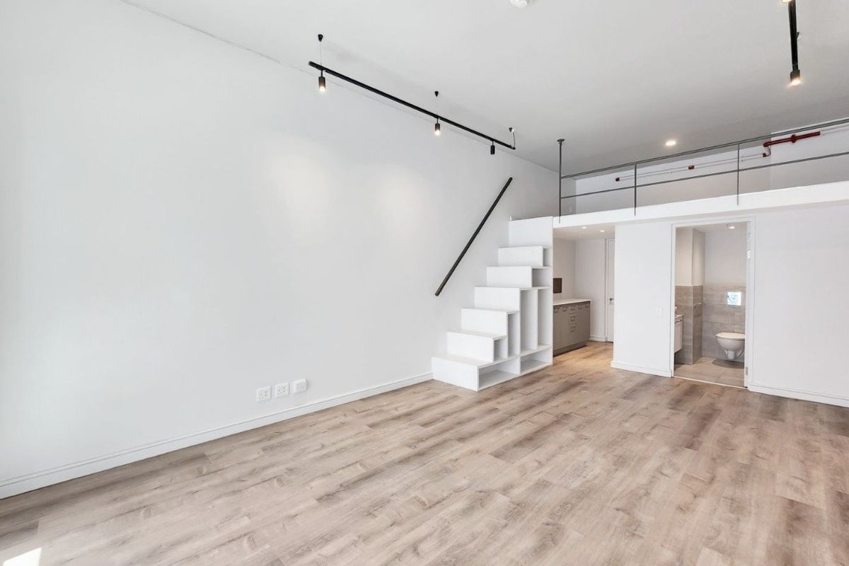 Modern open-plan apartment featuring light laminate flooring, a mezzanine level with metal railing, built-in staircase storage, and track lighting.