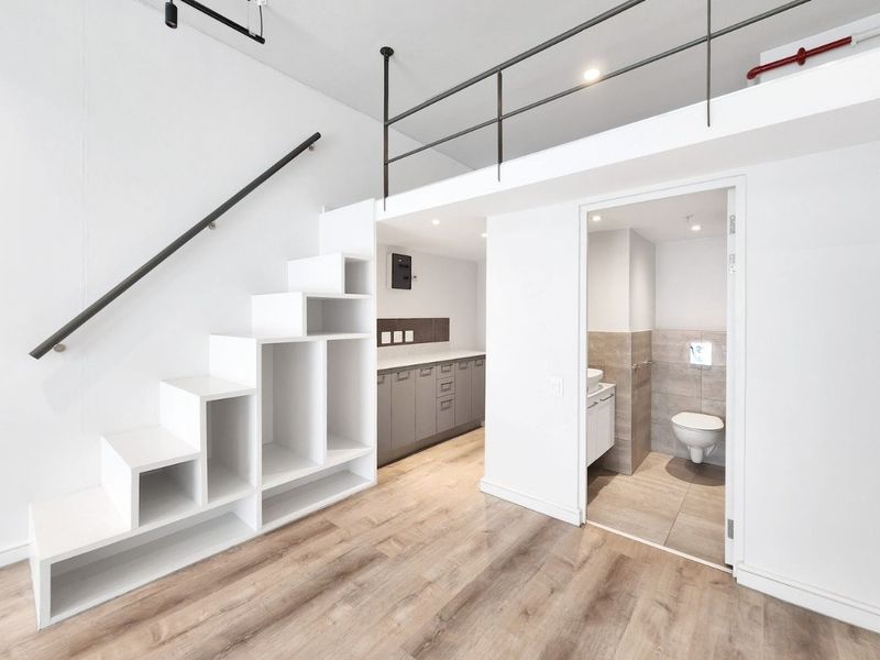 Modern open-plan apartment interior featuring a mezzanine level, built-in staircase storage, laminate flooring, and an adjacent bathroom with contemporary fixtures.