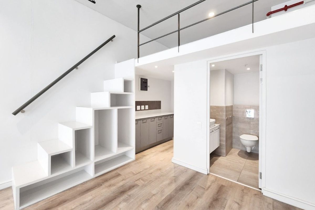 Modern open-plan apartment interior featuring a mezzanine level, built-in staircase storage, laminate flooring, and an adjacent bathroom with contemporary fixtures.