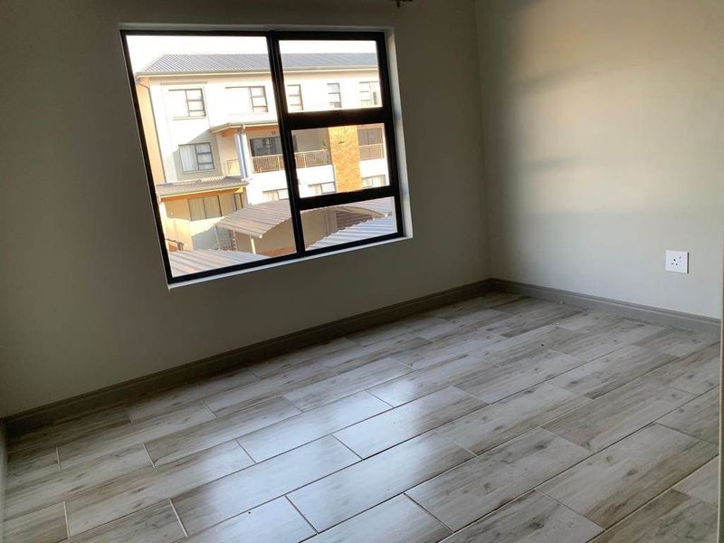 A bright, empty bedroom featuring modern wood-look ceramic floor tiles and a large aluminium-framed window with views of the surrounding complex.