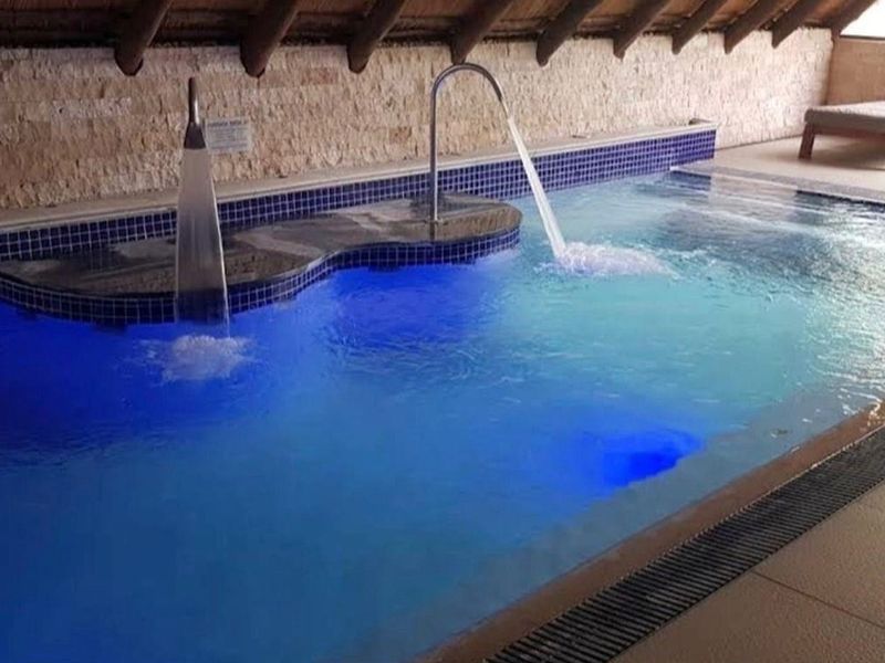 An indoor swimming pool area featuring stone-clad walls, a thatched roof, and integrated water features with blue ambient lighting.
