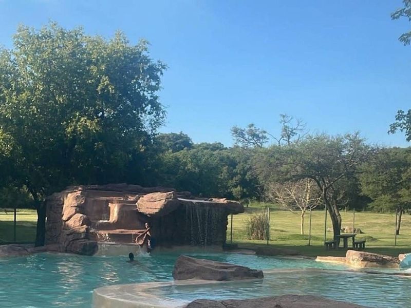 A landscaped outdoor pool area featuring a naturalistic rock waterfall, integrated water slide, and expansive surrounding green space.