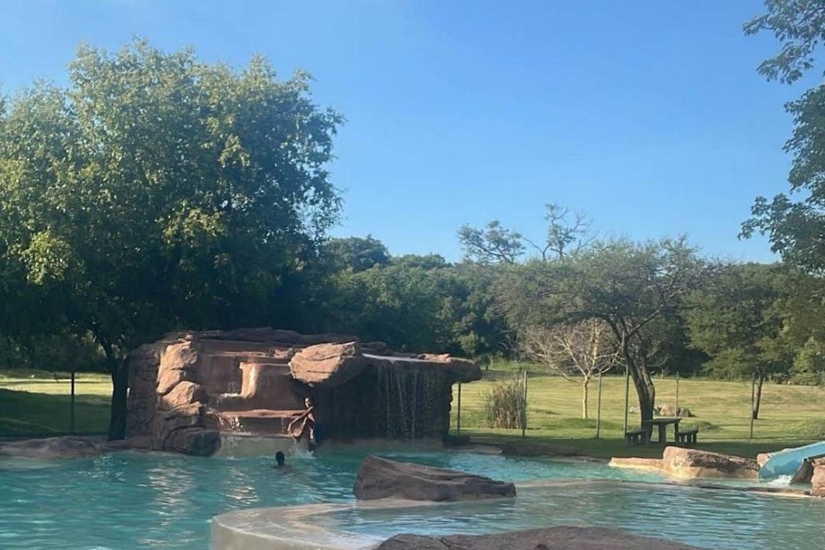A landscaped outdoor pool area featuring a naturalistic rock waterfall, integrated water slide, and expansive surrounding green space.