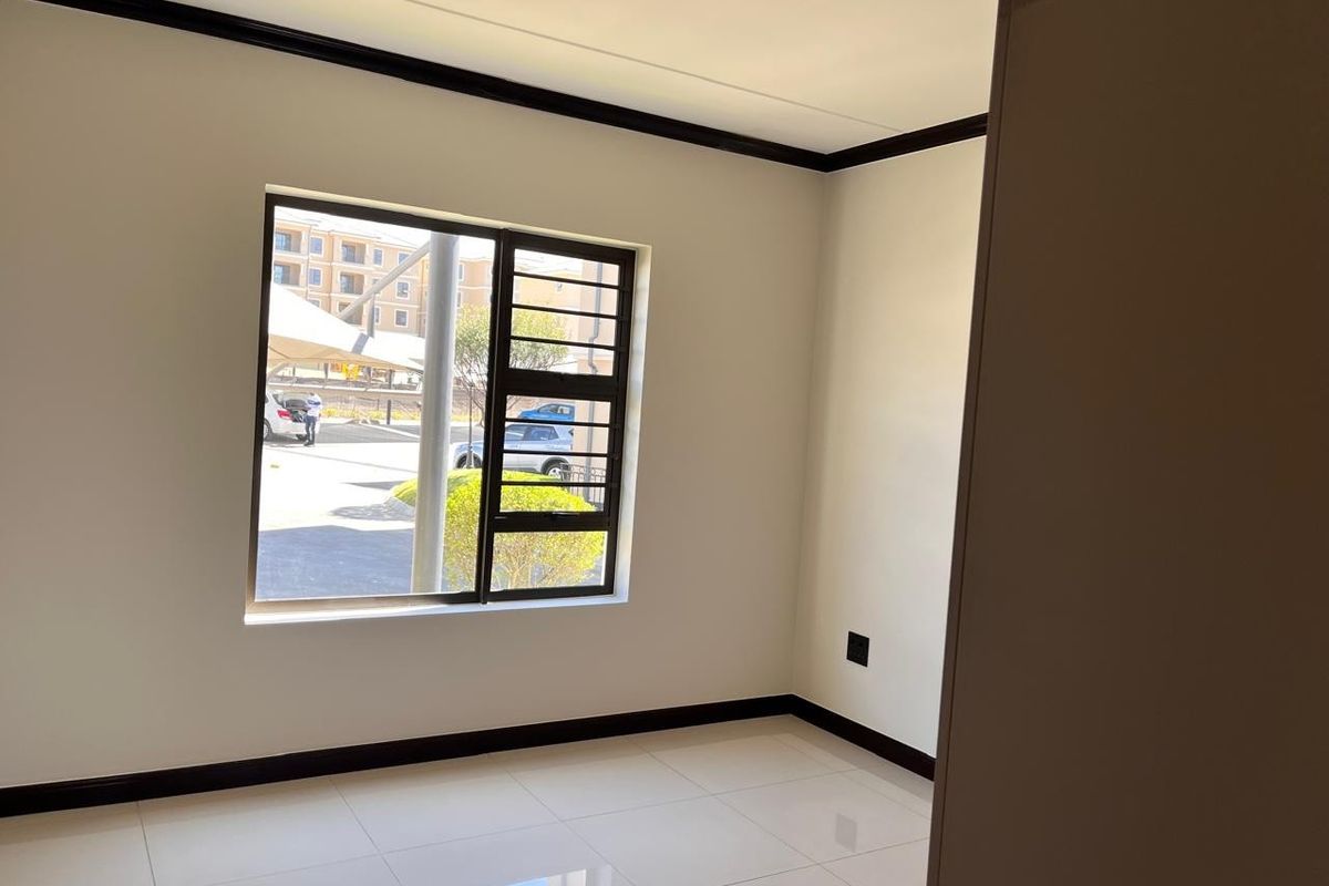 A bright, empty bedroom featuring neutral tiled flooring, dark skirting boards, and an aluminium window with integrated security bars.