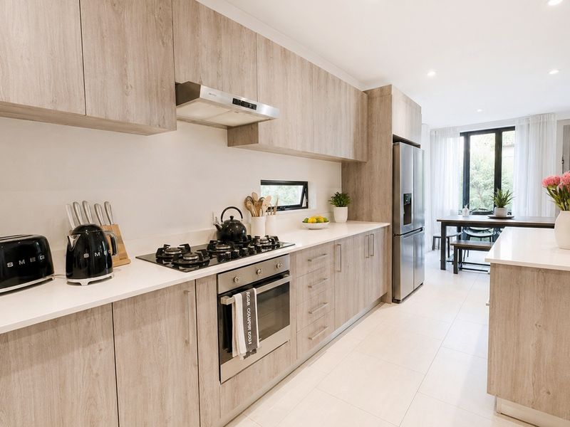 Modern open-plan kitchen featuring wood-grain cabinetry, stone benchtops, integrated appliances, and tiled flooring, flowing into a bright dining area.