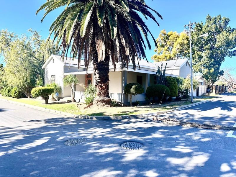 A charming corner-lot house featuring a covered veranda, gabled roof, and established garden with a prominent palm tree.
