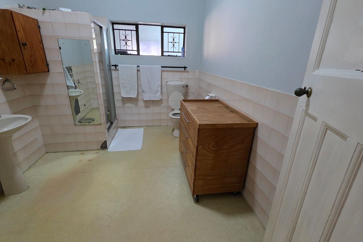 A dated bathroom featuring a pedestal basin, shower enclosure, and toilet, with original wall tiling and worn flooring throughout.