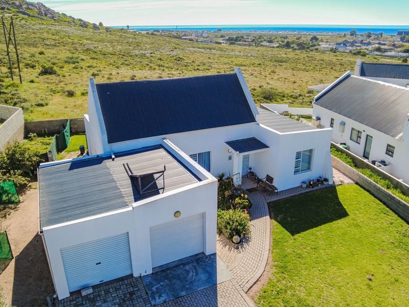 Aerial view of a white house with a corrugated metal roof, double garage, paved driveway, and a landscaped garden in a coastal setting.