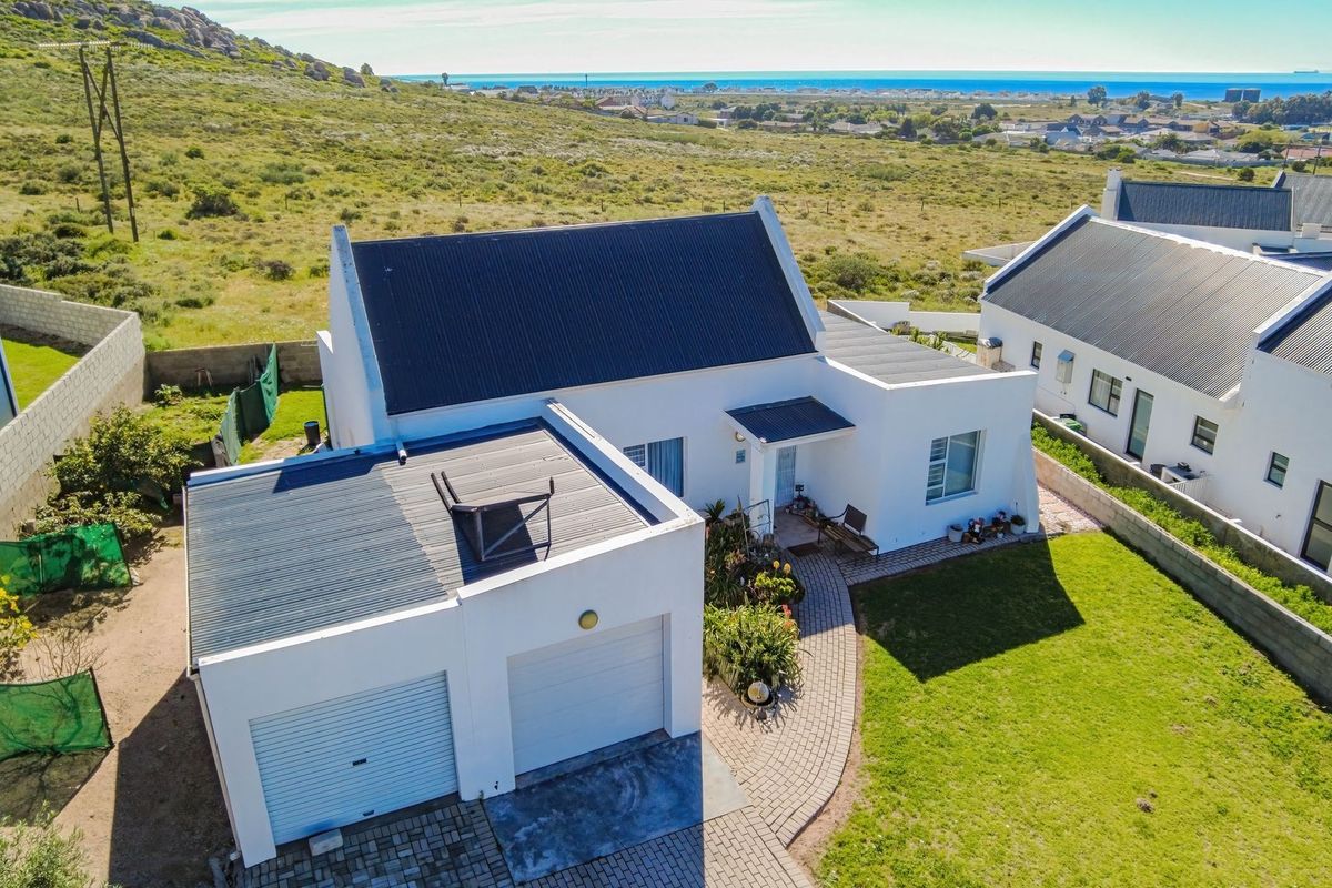 Aerial view of a white house with a corrugated metal roof, double garage, paved driveway, and a landscaped garden in a coastal setting.