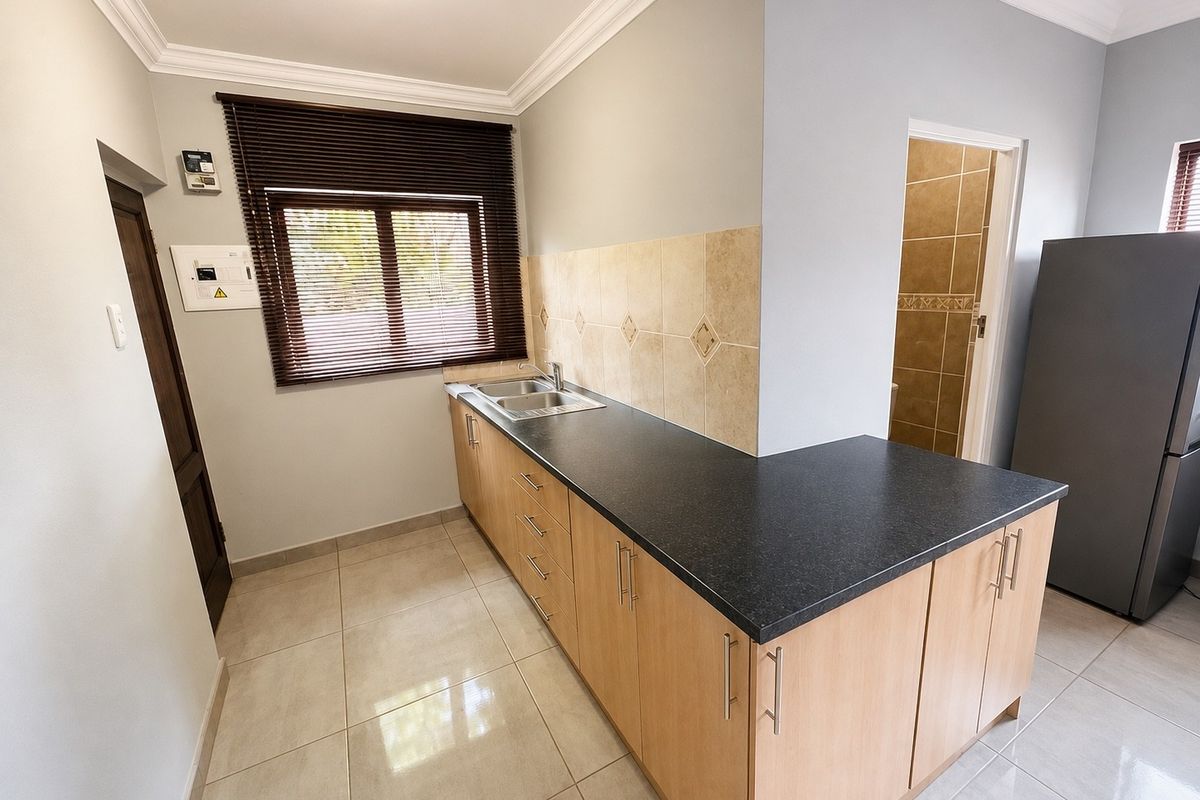 A functional kitchen featuring light wood cabinetry, dark laminate countertops, tiled backsplash, and neutral floor tiles, with an adjacent doorway leading to a bathroom.