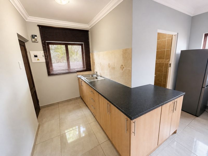A functional kitchen featuring light wood cabinetry, dark laminate countertops, tiled backsplash, and neutral floor tiles, with an adjacent doorway leading to a bathroom.