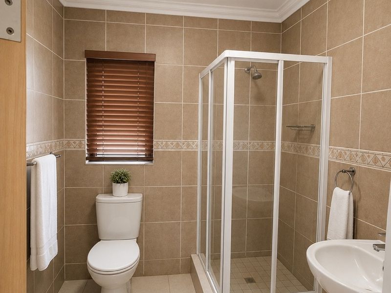 A clean, functional bathroom featuring a corner shower enclosure, pedestal basin, toilet, and neutral floor-to-ceiling wall tiling.