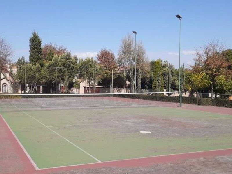 A private outdoor tennis court featuring perimeter fencing and floodlights, showing signs of wear on the playing surface.