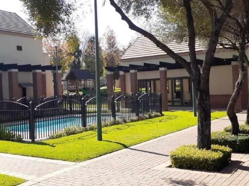A well-maintained outdoor pool area featuring a fenced swimming pool, brick paving, and covered pergola structures set within a landscaped garden.