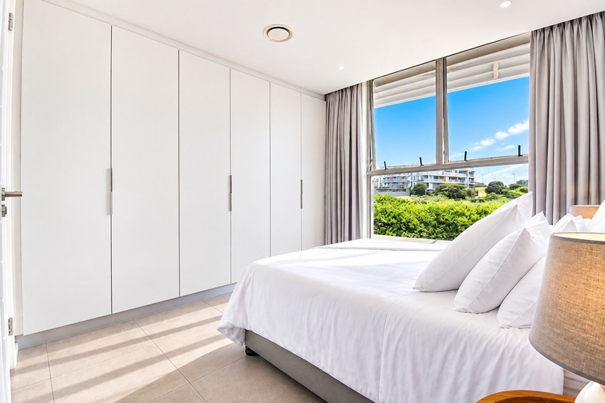 A bright, modern bedroom featuring extensive built-in wardrobes, tiled flooring, and a large window offering pleasant exterior views.