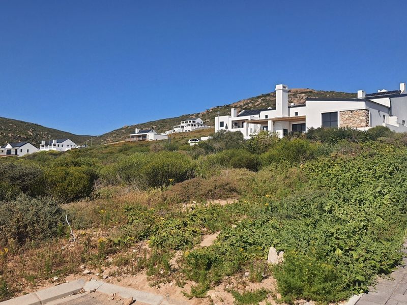 A vacant residential plot featuring natural coastal vegetation, a paved driveway edge, and views of established modern homes in the surrounding estate.