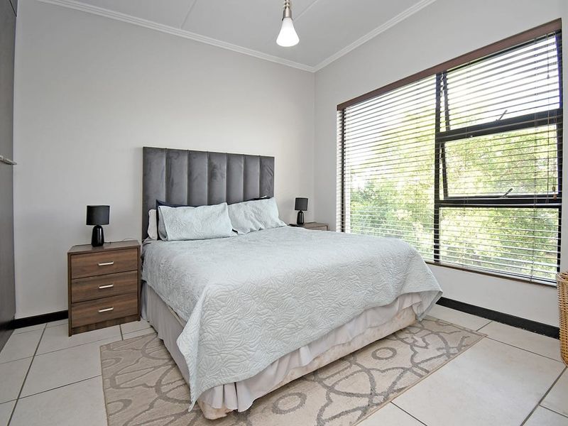 A well-lit bedroom featuring tiled flooring, built-in cupboards, and large windows with venetian blinds, offering a comfortable and functional living space.
