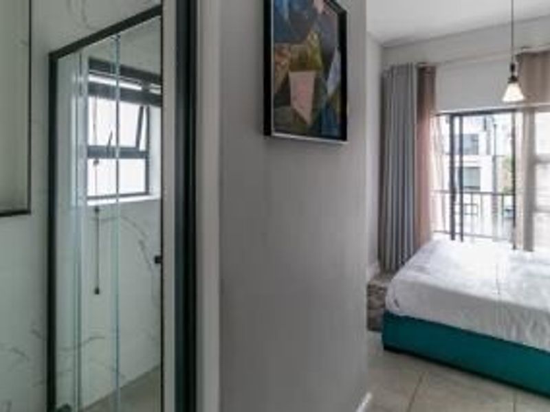 A modern bedroom featuring an en-suite glass shower enclosure with marble-look tiling and direct access to a balcony through sliding doors.