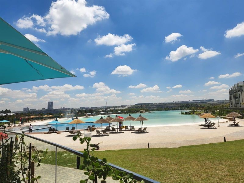 A view of a large communal swimming pool with a sandy beach area, landscaped lawns, and glass balustrade overlooking the estate.