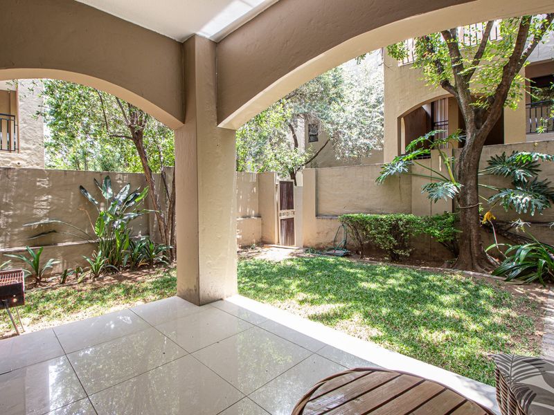 A covered patio area featuring tiled flooring, overlooking a private garden space with boundary walls and established trees.