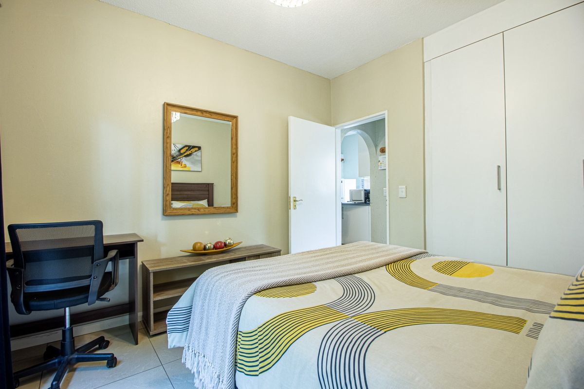 A functional bedroom featuring tiled flooring and a built-in wardrobe, with a view through the doorway into the adjacent kitchen area.
