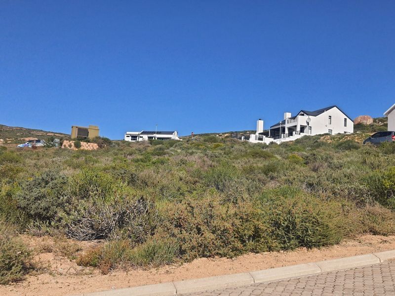 A view of a vacant residential plot featuring sloped terrain and native vegetation, situated in a developed coastal or hillside neighborhood.