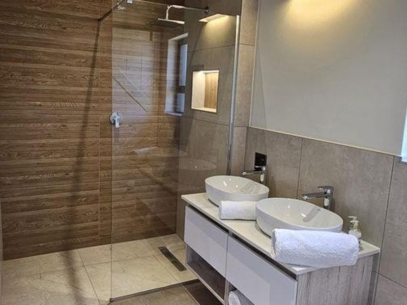 Modern bathroom featuring a double vanity, walk-in shower with glass screen, wall-mounted toilet, and stylish exposed concrete ceiling with integrated lighting.