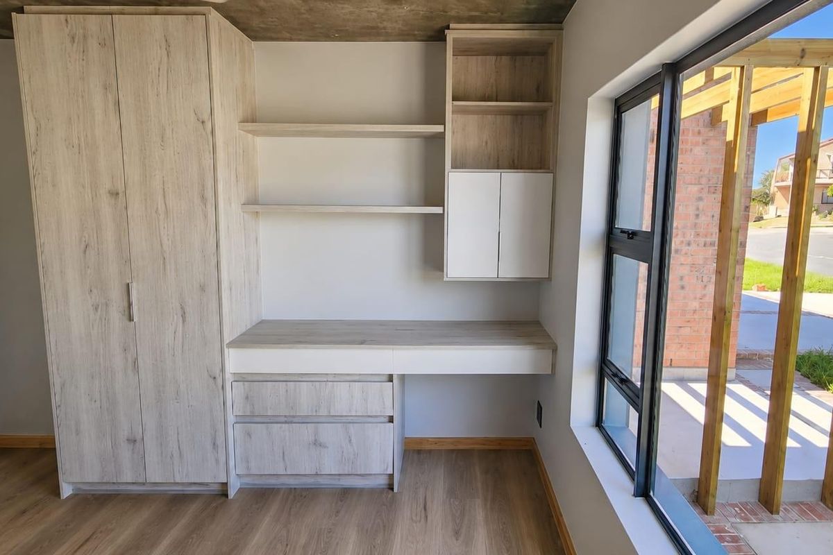 Modern bedroom featuring built-in cabinetry, desk, and shelving with laminate flooring, exposed concrete ceiling, and large windows overlooking the exterior patio.