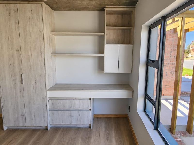Modern bedroom featuring built-in cabinetry, desk, and shelving with laminate flooring, exposed concrete ceiling, and large windows overlooking the exterior patio.