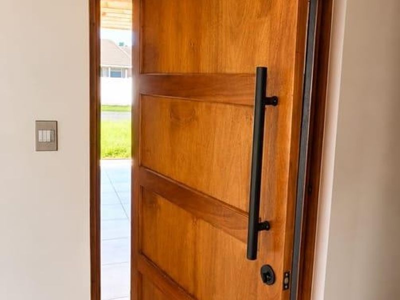 A modern entrance featuring a large timber pivot door with a sleek black handle, complemented by exposed wooden ceiling beams and light-toned tiled flooring.