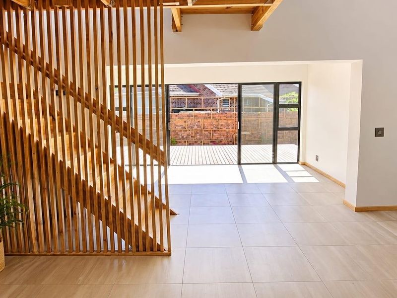Spacious open-plan living area featuring a modern timber slat staircase screen, exposed ceiling beams, tiled flooring, and seamless access to the rear patio.