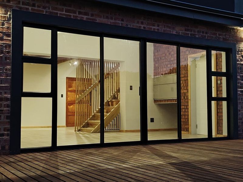 A modern outdoor timber deck featuring large sliding glass doors that provide a clear view into the bright, open-plan interior with a timber staircase.