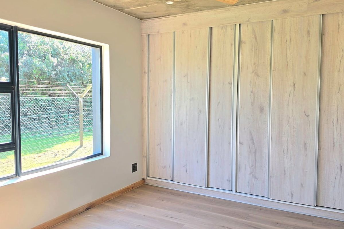 A bright, modern bedroom featuring light wood-grain built-in cupboards, exposed concrete ceilings, and large windows overlooking the garden.