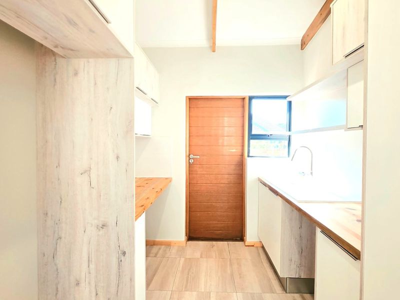 A clean, modern scullery featuring light wood-look cabinetry, timber countertops, a stainless steel sink, and tiled flooring with a wooden door.