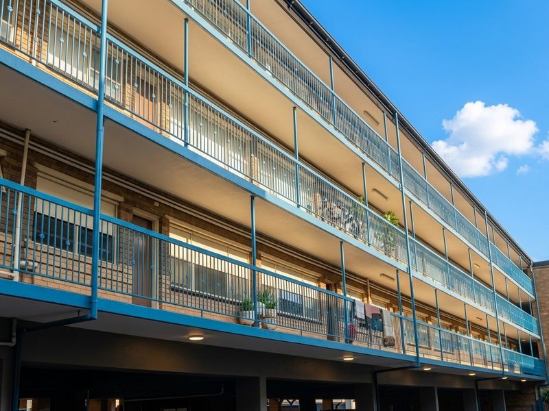 A multi-story apartment building exterior featuring brick facade, tiered balconies with metal railings, and an integrated ground-level undercover parking area.
