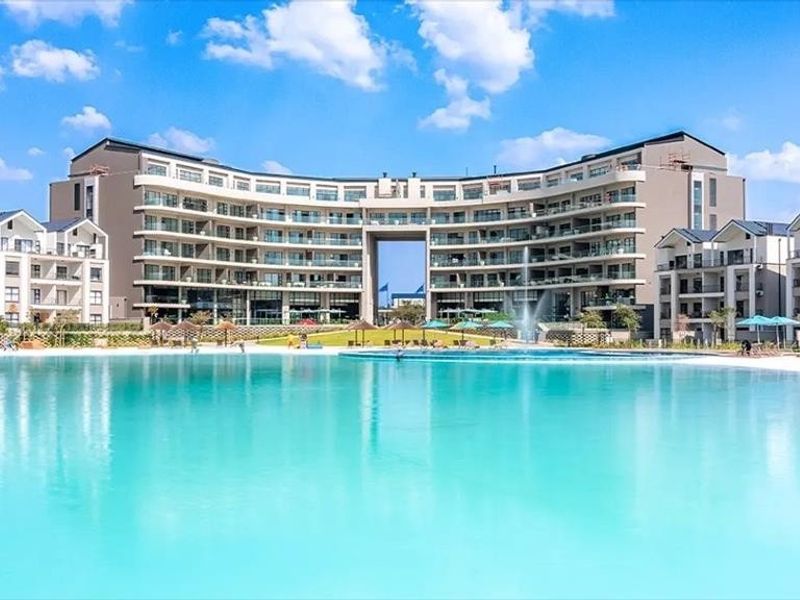 A large, clear swimming pool with a central fountain, set against a modern apartment complex with multiple balconies and landscaped grounds.