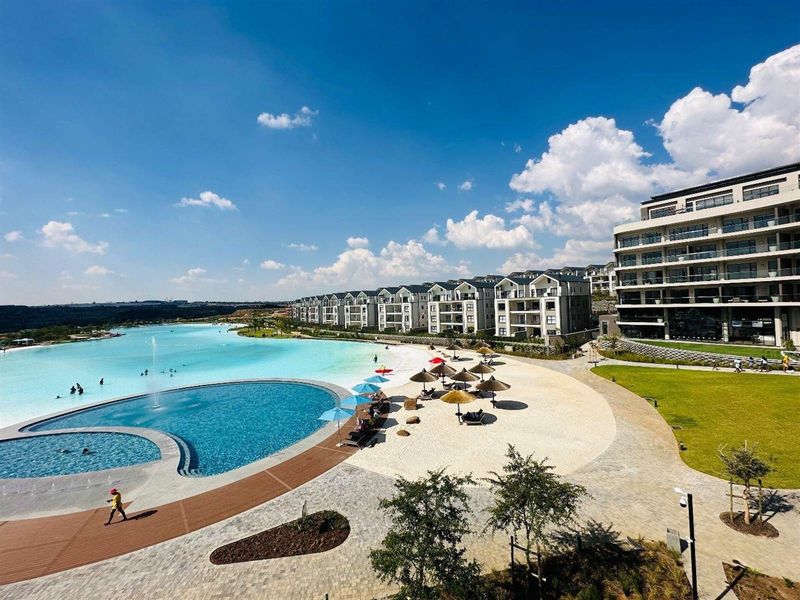 Aerial view of a luxury apartment complex featuring a large swimming pool, man-made beach area, and well-maintained landscaped grounds.