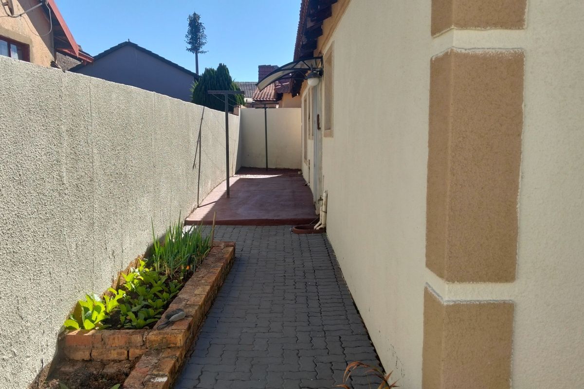 A narrow rear exterior side passage featuring a paved walkway, a small brick planter box, and a concrete patio area.