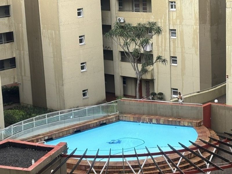 An elevated view of a communal swimming pool area featuring a paved surround, metal safety railings, and a pergola structure set against apartment building facades.