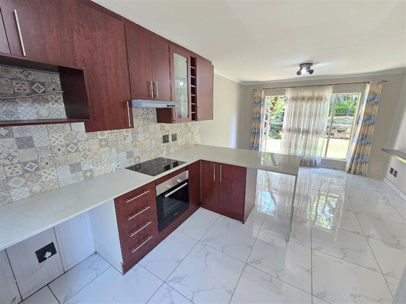 Open-plan kitchen featuring dark wood cabinetry, stone countertops, built-in oven and hob, and modern tiled flooring, leading to a bright living area.