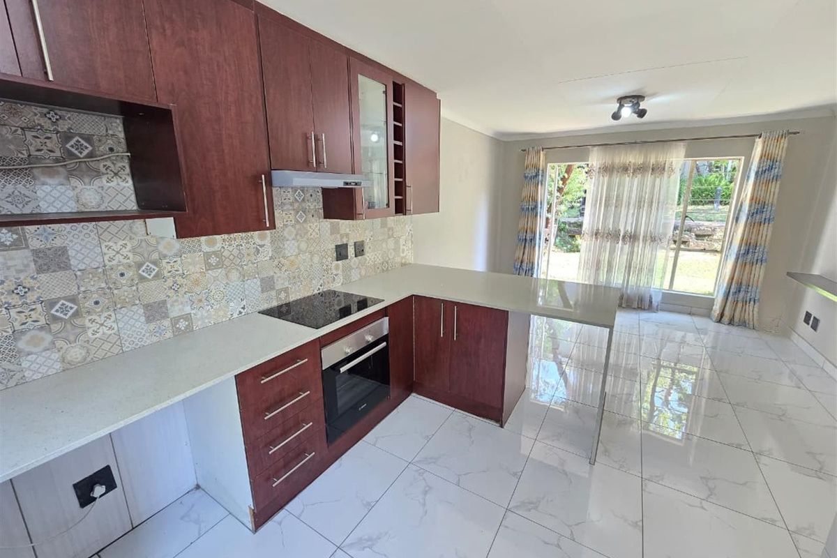 Open-plan kitchen featuring dark wood cabinetry, stone countertops, built-in oven and hob, and modern tiled flooring, leading to a bright living area.