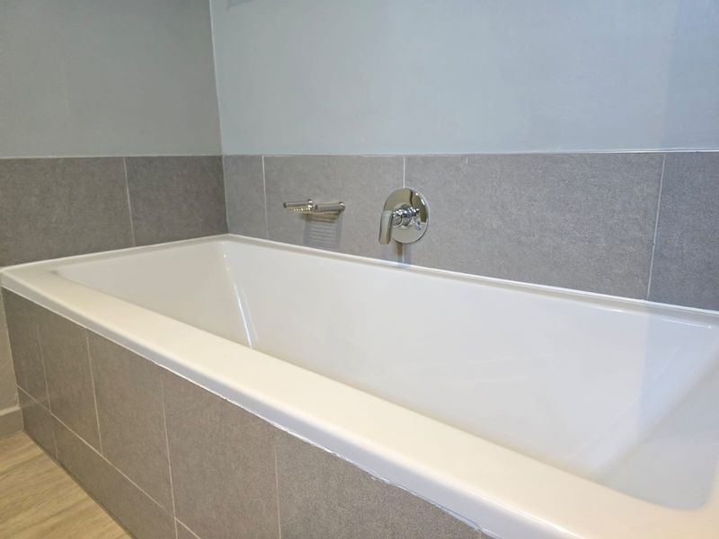 A clean, modern bathroom featuring a built-in bathtub with neutral grey wall tiles and polished chrome fixtures.