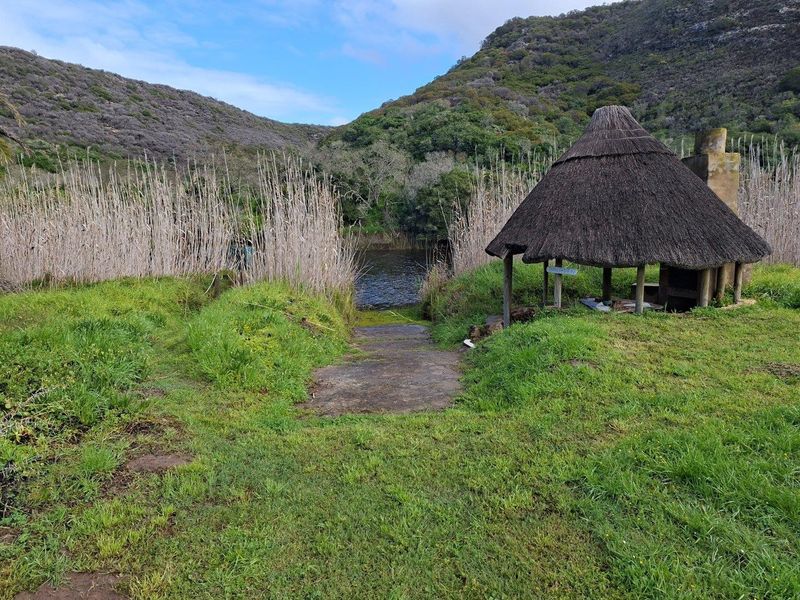 Outdoor recreational area featuring a traditional thatched lapa with a built-in braai, overlooking a natural water body with a concrete boat launch ramp.